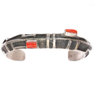 May include: A cuff bracelet featuring a dark, oxidized metal finish with a textured, geometric surface. Two rectangular, vibrant orange stones are set into the metal, one positioned centrally and one toward the right side of the band.