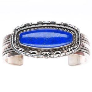 May include: A silver cuff bracelet featuring a central, elongated, rectangular lapis lazuli stone with gold-coloured flecks. The stone is set within a decorative silver frame with scalloped edges and twisted wire details, flanked by triple-wire bands.