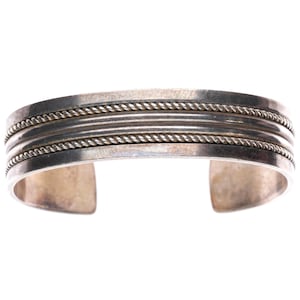 May include: A silver cuff bracelet featuring a smooth, polished center band flanked by two thin, twisted rope-style textured bands on the top and bottom edges. The metal has a metallic sheen with subtle signs of natural oxidation.