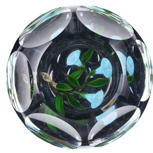 May include: A clear glass paperweight with a floral design. The interior features light blue flowers and green leaves. The exterior has a series of oval-shaped cutouts. The paperweight is round and sits on a white background.