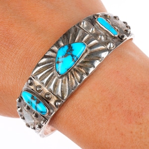 May include: A silver cuff bracelet featuring three turquoise stones. The bracelet has a sunburst design around the central stone and is decorated with small, round studs. The turquoise stones are a bright blue colour.