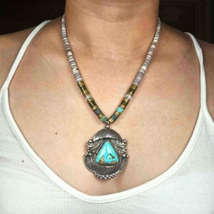 Puede incluir: Un collar con colgante de plata con una piedra turquesa. El collar tiene un hilo de cuentas con cuentas blancas, grises y verdes. El colgante es un diseño ornamentado con una piedra turquesa triangular.