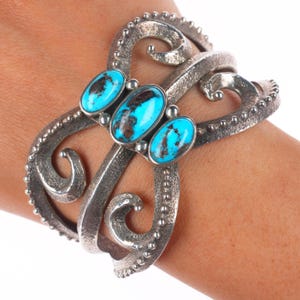 May include: A silver cuff bracelet featuring three oval turquoise stones. The bracelet has an open design with swirling, textured metalwork and a dotted border. The turquoise stones are a vibrant blue with brown matrix.