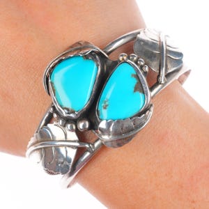 Puede incluir: Brazalete de plata con dos piedras turquesas brillantes. El brazalete tiene un diseño de hojas y cuentas, con las piedras turquesas engastadas en un marco decorativo. El brazalete mide aproximadamente 6,35 cm de ancho.