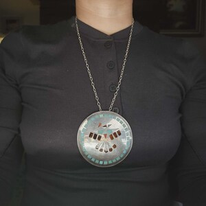 May include: A large circular silver pendant necklace featuring a central inlaid mosaic design of a bird with wings spread. The inlay consists of turquoise, coral, and dark stone pieces, surrounded by a border of small square turquoise tiles.
