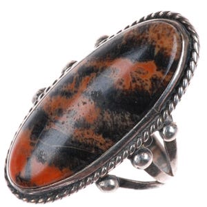 May include: A silver ring featuring an oval-shaped stone with a unique pattern of black, gray, and reddish-brown hues. The stone is set in a silver bezel with a rope-like border and decorative silver accents. A statement piece.