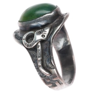 May include: A silver ring featuring a smooth, dark green oval stone set in a raised bezel. The band is decorated with an embossed snake motif on the side and a small geometric swastika symbol engraved near the bottom of the metal shank.