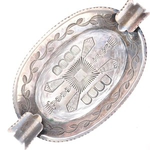 Puede incluir: Cenicero ovalado de plata con un diseño grabado detallado. El cenicero presenta un patrón geométrico central con líneas radiales y formas estilizadas. El borde está decorado con un patrón de hojas repetido. Dos apoyos para ceniceros están en lados opuestos.