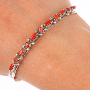 May include: A silver bracelet with vibrant red coral stones. The bracelet features a series of oval-shaped coral pieces set in individual silver bezels, alternating with small silver beads. The design is set on a thin, curved silver band.
