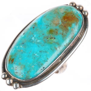 May include: An oval-shaped ring featuring a large, polished turquoise stone with natural brown matrix inclusions. The stone is set in a smooth silver bezel, accented by three small silver spheres at both the top and bottom ends of the setting.