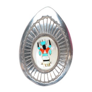 May include: A teardrop-shaped silver brooch featuring a central oval inlay of mother-of-pearl, turquoise, coral, and black stone arranged in a stylized figure. The silver frame has a radiating sunburst pattern with etched lines around the edge.