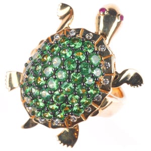 May include: A gold turtle ring with a shell of green gemstones. The turtle's head has two small red gemstones for eyes. Small white gemstones are set around the shell's edge. A unique piece of jewellery.
