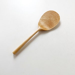 May include: A wooden rice paddle with a textured surface. The paddle is light brown in color and has a long handle.
