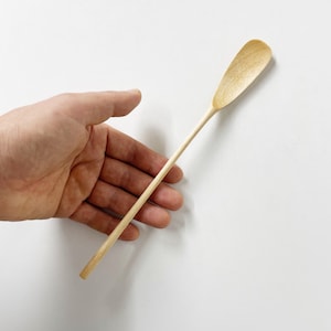 Puede incluir: Una cuchara de madera larga y delgada con un extremo redondeado ligeramente curvado. La cuchara es de color marrón claro y se sostiene en una mano.