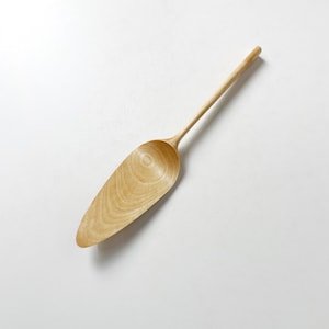 May include: A wooden serving spoon with a long, slender handle and a wide, rounded scoop. The spoon is made of light-colored wood and has a natural finish.