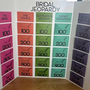 May include: A handmade game board titled "Bridal Jeopardy" with categories like "The Bride" and "The Couple." Each category has a series of envelopes with point values from 100 to 500. The board is made of cardboard and is set on a wooden surface.
