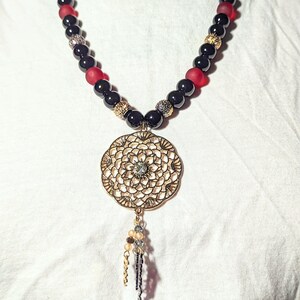 May include: A black and red beaded necklace with a gold filigree pendant featuring a crystal in the center. The pendant has a dangling chain with small beads.