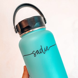 May include: A turquoise water bottle with a black handle and lid. The bottle has a black vinyl sticker with the name "sadie" written in cursive.