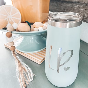 May include: A light blue tumbler with a silver rim and lid, featuring the initials "LP" and a heart. The tumbler is on a gray surface, next to a decorative beaded garland and a yellow vase with flowers.