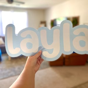 May include: A light blue sign with the name "Layla" in white, bubble-style letters. The sign is held up in front of a blurred background of a living room. The sign is a decorative item.