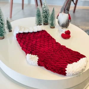 Peut inclure: Une chaussette de Noël rouge et blanche en tricot avec une texture moelleuse. La chaussette est posée sur une surface blanche.
