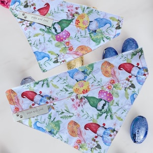 May include: Two white dog bandanas with a white background and a colourful pattern of gnomes, flowers, and Easter eggs. The bandanas have a silver tag that says "pops and fred" with paw prints.