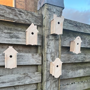 Puede incluir: Cinco casas de pájaros de madera de diferentes formas y tamaños están montadas en una valla de madera. Las casas de pájaros están pintadas en un color madera claro y tienen pequeñas aberturas para que los pájaros puedan entrar.