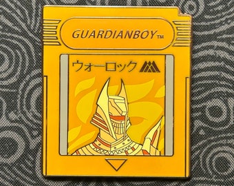 May include: A yellow and gold enamel pin shaped like a video game cartridge. The pin features a stylized image of a golden warrior with a helmet and a red visor. The text "GUARDIANBOY™" is printed on the top of the cartridge and the text "ウォーロック" is printed on the front of the cartridge.
