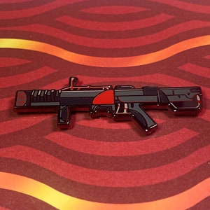 May include: A black and red enamel pin of a futuristic-looking gun. The gun has a red barrel and a black handle.