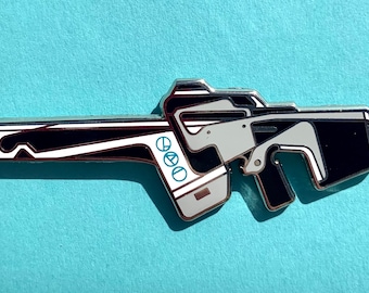 May include: A silver-colored metal pin shaped like a futuristic gun with black and white details. The gun has a white stripe running down the barrel and a circular design on the side.