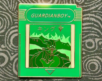 May include: Green and gold enamel pin featuring a video game cartridge design. The pin depicts a green monster with a red eye and the text "GUARDIANBOY" and "キング".