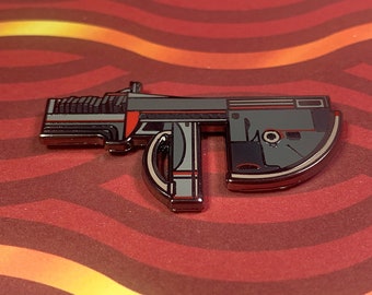May include: A silver and black enamel pin of a futuristic weapon with red accents. The pin is shaped like a gun with a curved barrel and a handle.
