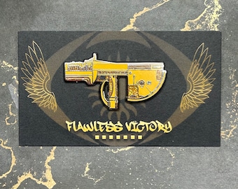 May include: Gold-colored metal pin featuring a detailed design of a futuristic weapon with a textured surface. The pin is displayed on a black background with the text "Flawless Victory" printed below the weapon.