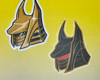 May include: Two vinyl stickers featuring stylized illustrations of anubis, the Egyptian god of the dead. The first sticker is gold with black stripes and the second sticker is black with gold stripes.
