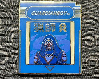May include: A blue and gold enamel pin featuring a stylized character design. The pin has the text "GUARDIANBOY" at the top and Japanese characters in the center. The character is depicted in shades of blue and purple.