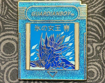 May include: A blue and gold enamel pin featuring a blue and white illustration of a creature with a crown and the text "氷の女王" in Japanese. The pin is shaped like a video game cartridge and has the text "GUARDIANBOY" on the top.