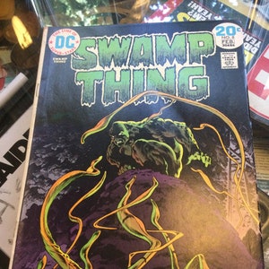 May include: A vintage comic book titled "Swamp Thing" featuring a green swamp creature on a dark background. The cover has the DC Comics logo and the issue number 8, dated February. The artwork includes vibrant green and orange plant-like tendrils.
