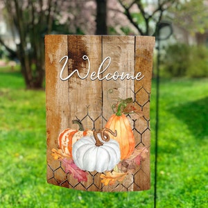 May include: A wooden sign with the word "Welcome" in white script. The sign is decorated with a white pumpkin, an orange pumpkin, and a striped pumpkin. The pumpkins are surrounded by fall leaves and a wire fence.