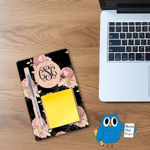 May include: A black notepad with a floral design and a monogram, a yellow sticky note, and a pen. The notepad has a pink floral design with a monogram in the center. A laptop is in the background.