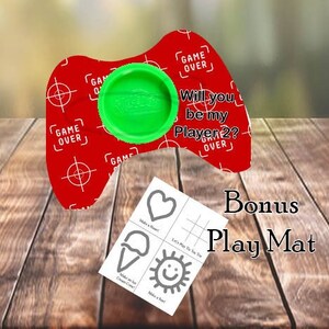 May include: A red and white game controller shaped dough holder with a green lid. The holder has the text "Game Over" on it. The lid has the text "Will you be my Player 2?" on it. A bonus play mat with a heart, ice cream cone, and sun design is included.