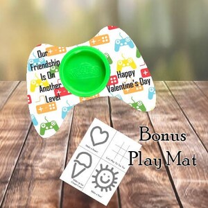 Könnte beinhalten: Ein weißer und grüner Gamecontroller-förmiger Teigbehälter mit einer grünen Mitte zum Halten von Teig. Der Controller hat ein farbenfrohes Design mit dem Text "Our Friendship Is On Another Level" und "Happy Valentine's Day". Eine Bonus-Spielmatte mit einem Herz-, Eiswaffel- und Sonnendesign ist enthalten.