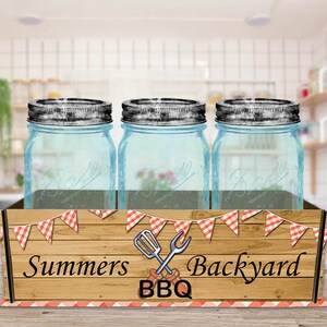 May include: Three clear glass jars with metal lids are displayed in a wooden crate with a red and white checkered pattern. The crate has the text "Summers Backyard BBQ" printed on it.