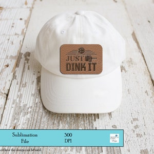 K&ouml;nnte beinhalten: Wei&szlig;e Baseballkappe mit einem braunen Lederpatch, auf dem "JUST DINK IT" steht, mit einer Grafik eines Basketballkorbs und eines Balls.