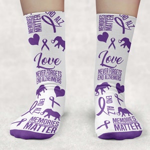 Puede incluir: Calcetines morados y blancos con el texto "Memories Matter", "End Alz", "Love" y "Never Forgets End Alzheimers". Los calcetines también presentan una cinta morada y un gráfico de elefante.