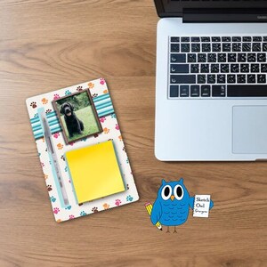 Può includere: Un organizzatore da scrivania colorato con una foto di un cane nero, un post-it giallo, una penna e disegni di impronte di zampe. L'organizzatore è su una superficie di legno.
