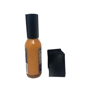 Puede incluir: Una botella de vidrio transparente con una tapa negra llena de salsa picante naranja. La botella está sobre una superficie blanca junto a una pila de etiquetas negras.