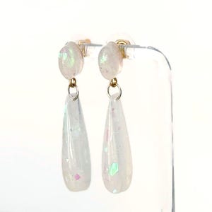 May include: A pair of dangle earrings with a pearlescent finish. Each earring features a small, round stud with a gold-colored post, connected to a teardrop-shaped charm. The charms are a translucent white with iridescent glitter accents.