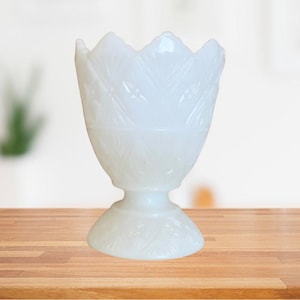 May include: A white, opaque glass vase with a fluted design. The vase has a wide, round base and a narrow, flared top.