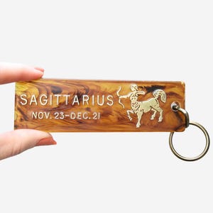 May include: A rectangular keychain with a brown and amber marbled pattern. The keychain features the word "SAGITTARIUS" and the dates "NOV. 23-DEC. 21" in white text. A gold-colored Sagittarius symbol and a gold-colored keyring are also present.