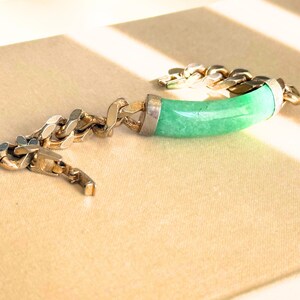May include: A silver-toned chain bracelet with a curved, light green jade centerpiece. The jade is set in a silver metal frame. The bracelet is displayed on a beige surface.